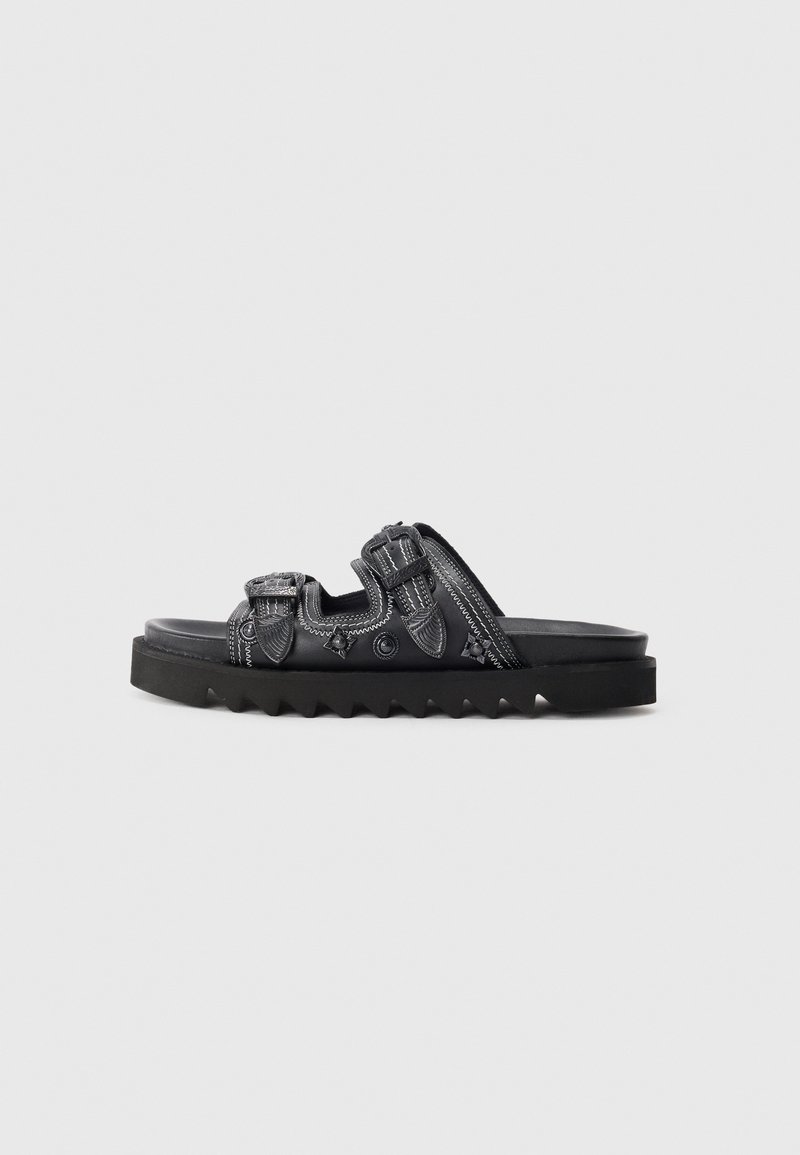 Black sandal with three adjustable straps, white stitching, decorative metal details, and thick jagged sole on a white background.