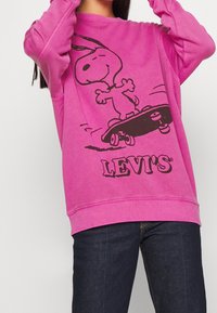 Sudadera rosa con un gráfico negro de Snoopy en una patineta, con el logo "LEVI'S" impreso debajo del gráfico. El material parece suave y texturizado.