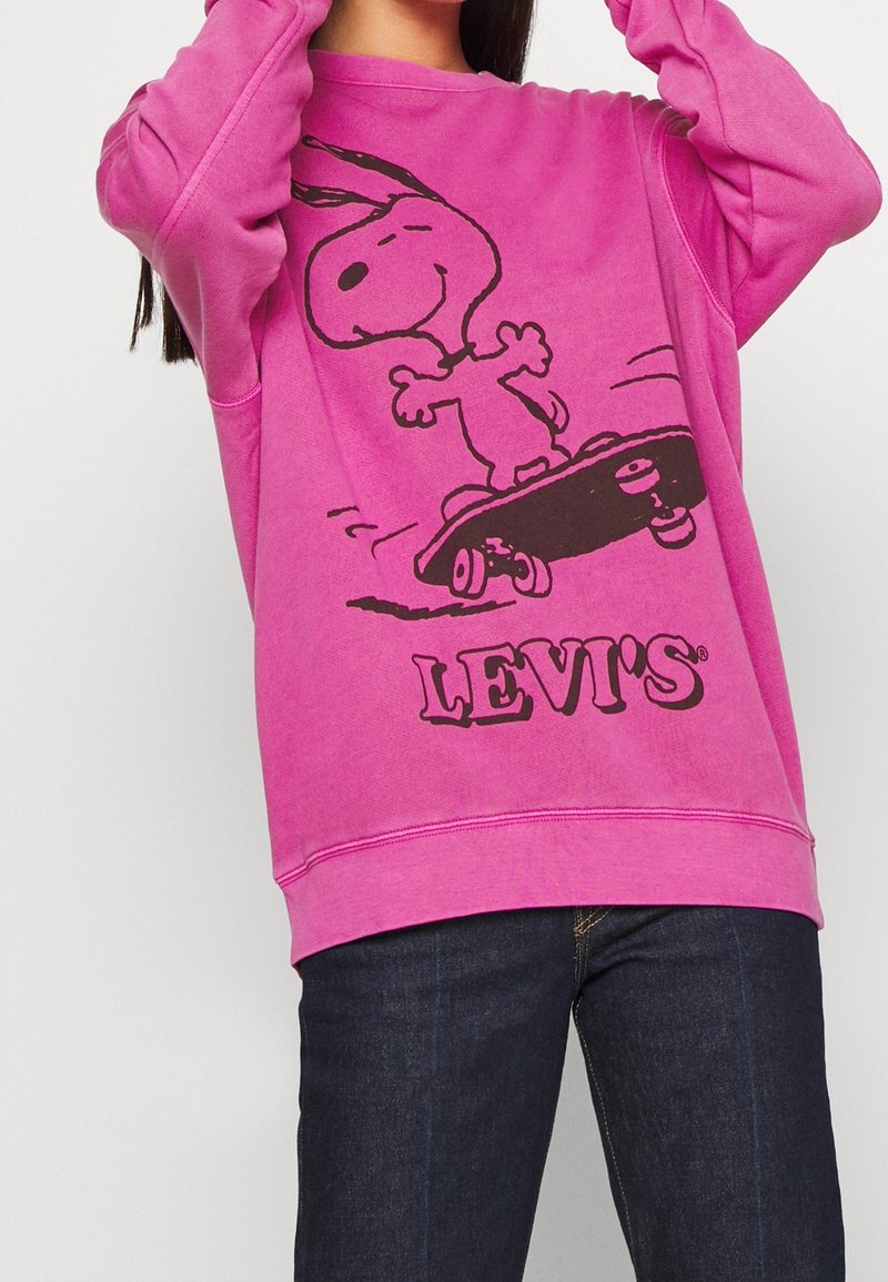 Sudadera rosa con un gráfico negro de Snoopy en una patineta, con el logo "LEVI'S" impreso debajo del gráfico. El material parece suave y texturizado.