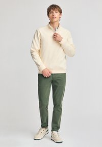 Suéter de cremallera en color crema, pantalones verdes y zapatillas beige. El suéter presenta una textura suave; los pantalones tienen un acabado liso y un corte ajustado.