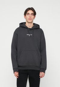 Pelēks kokvilnas kapuce ar priekšējo kabatu, aukliņu kapuci un baltu "ADIDAS ORIGINALS" logotipu uz krūšu. Brīvs piegriezums.