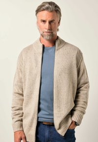 Bežový pletený kardigan s vysokým límcem, žebrovanými manžetami a strukturovaným povrchem. Nosený přes světle modrou košili, v kombinaci s tmavými džíny.