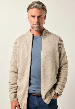 Cárdigan de punto beige con cuello alto, puños acanalados y un acabado texturizado. Lleva encima de una camisa azul clara, combinada con jeans oscuros.