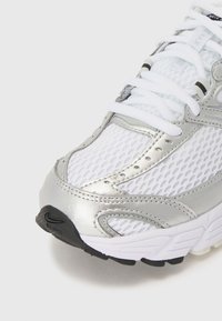 Chaussure de sport blanche et argentée avec tige en mesh, lacets blancs et semelle extérieure noire visible à l'avant et sur le côté.
