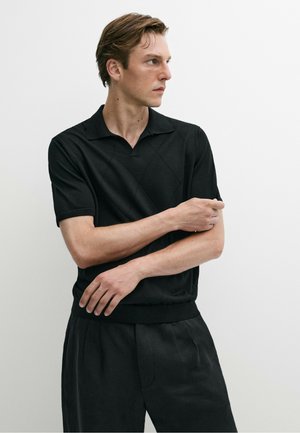 Mann mit heller Haut und braunen Haaren, der ein schwarzes kurzärmeliges Hemd mit Kragen und schwarze Hose trägt, steht vor einem schlichten weißen Hintergrund.