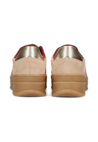 Vista posteriore di scarpe con una tomaia in suede color beige, dettagli metallici dorati e una suola spessa marrone. Presentano una texture liscia e dettagli di colore a contrasto.