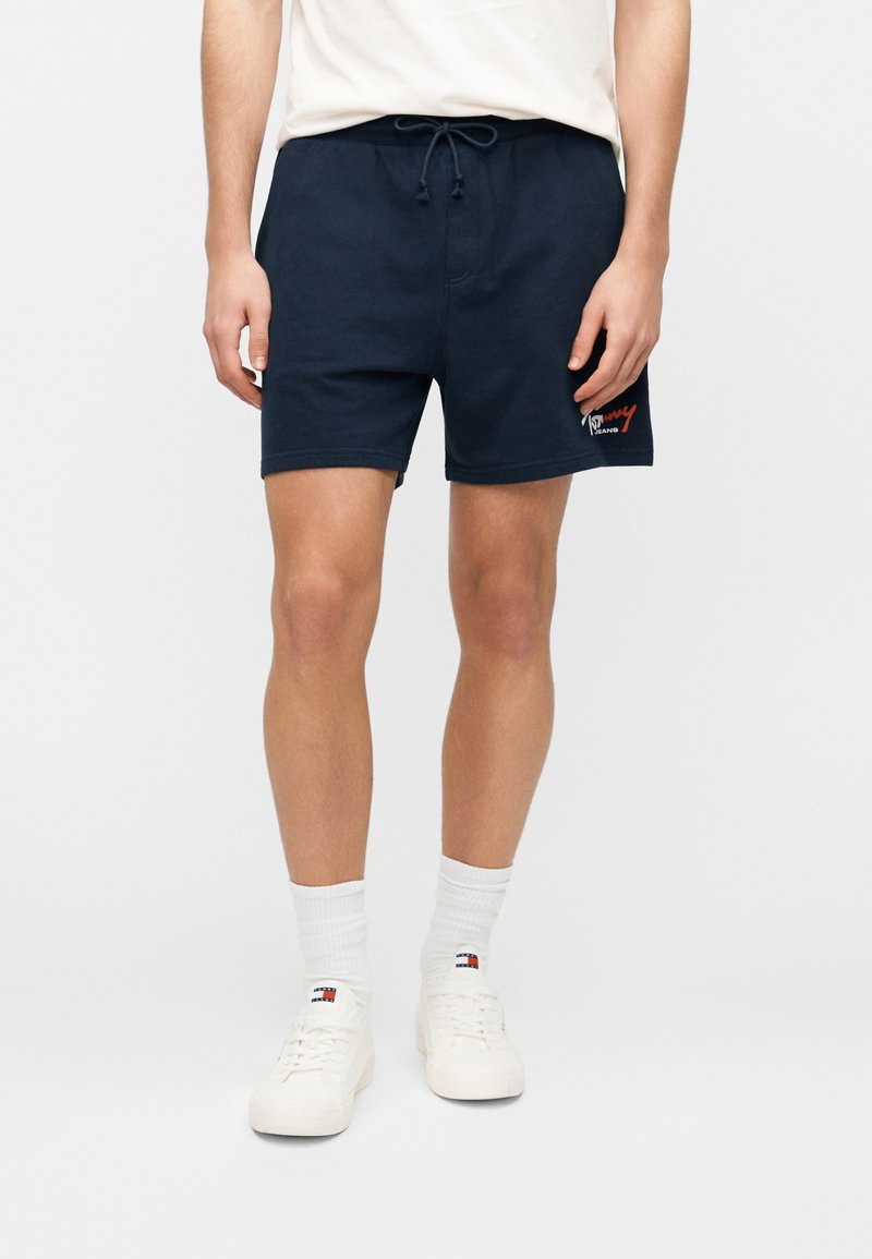 Μπλε σκούρο βαμβακερό σορτς με ελαστική μέση και κορδόνι, με λογότυπο σε κόκκινο στο αριστερό μηρό. Φοράνε με λευκά αθλητικά παπούτσια και κάλτσες.