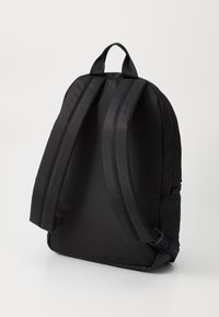 Sac à dos en tissu noir avec des bretelles ajustables, une poignée supérieure et un design minimalist. Texture lisse avec un branding subtil sur la sangle.