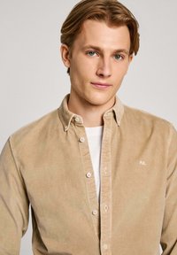 Beige corduroy shirt met een button-down kraag, voorzien van een wit logo en zichtbare knopen, gelaagd over een wit T-shirt.