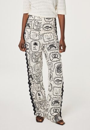 Vrouw die witte broek met hoge taille draagt, met zwart abstracte lijstafbeeldingen en gerimpelde zwarte zijstrepen, gecombineerd met een witte gebreide top en sandalen.