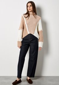 Sweter dzianinowy z golfem w beżowym i białym kolorze, z diagonalnym wzorem i teksturowanymi rękawami; zestawiony z ciemnymi spodniami o szerokim kroju.