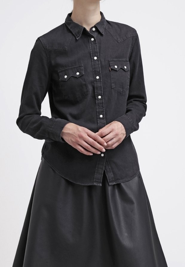 Personne portant une chemise noire boutonnée avec deux poches poitrine et une jupe noire, debout, les mains jointes devant.