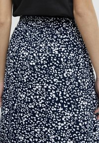 Žena v námořnické modré sukni s bílým abstraktním leopardím vzorem a černém topu, zobrazená zezadu od pasu dolů.
