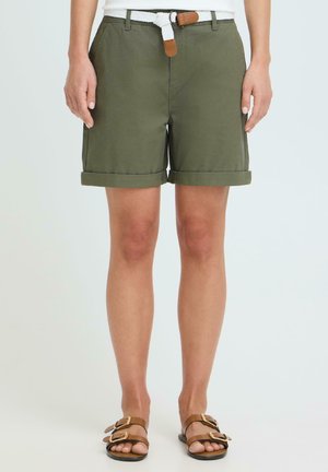 Osoba nosząca oliwkowozielone szorty z podwiniętymi nogawkami, z białym plecionym paskiem i brązowymi sandałami na podwójny pasek, stojąca na jednolitym tle.