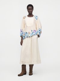 Femme portant une blouse ample crème avec une broderie florale bleue et violette, une jupe longue assortie, et des bottes marron mi-mollet, debout sur fond blanc.