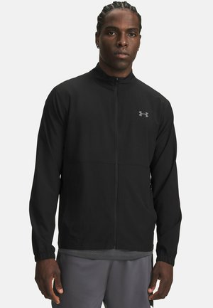 Mann mit geflochtenen Haaren trägt eine schwarze Under Armour Jacke mit Reißverschluss und graue Hose, steht vor weißem Hintergrund.