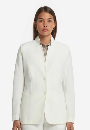Blazer bianco su misura con colletto alto, chiusura a due bottoni e tasche laterali. Tessuto testurizzato con una finitura liscia. Camicia a righe sotto.