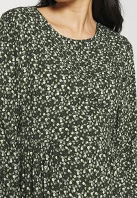 Robe florale en tissu vert foncé, avec un décolleté en scoop, des manches longues et des motifs floraux crème dispersés, avec des accents froncés à la taille.