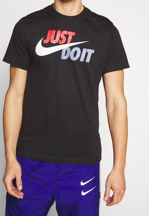 Person, die ein schwarzes Nike T-Shirt mit rotem und blauem "JUST DO IT" Text und zwei weißen Nike-Swooshes auf blauen Shorts trägt.