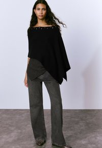 Poncho negro de canalé con diseño off-shoulder, decorado con tachuelas metálicas. Combinado con jeans de talle alto y pernera amplia en gris oscuro y zapatos con tachuelas.