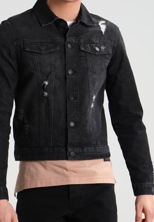 Homme portant une veste en jean noire usée sur une chemise beige, avec un pantalon noir, sur un fond uni clair.