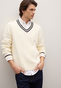 Roomkleurige kabelgebreide trui met een V-hals en marineblauwe strepen, gedragen over een wit overhemd met kraag, gecombineerd met donkere jeans.