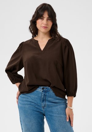 Femme aux cheveux foncés portant un chemisier marron ample à manches trois-quarts et un jean bleu sur un fond uni clair.