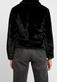 Svart fuskpälsjacka med mjuk textur, med en krage och långa ärmar, tillsammans med grå högmidjade jeans.