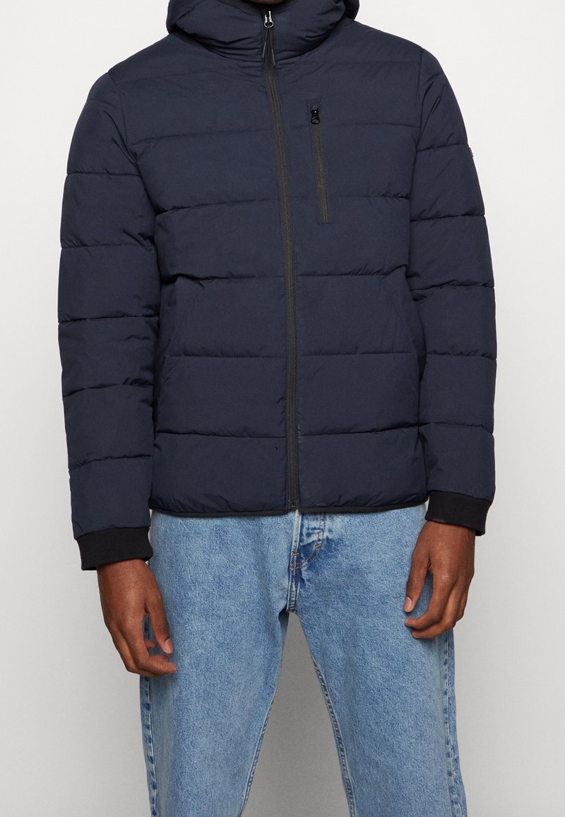 Mężczyzna ubrany w granatową pikowaną kurtkę puchową z przednim zamkiem błyskawicznym i zapinaną na zamek kieszenią na piersi, w połączeniu z jasnoniebieskimi dżinsami.