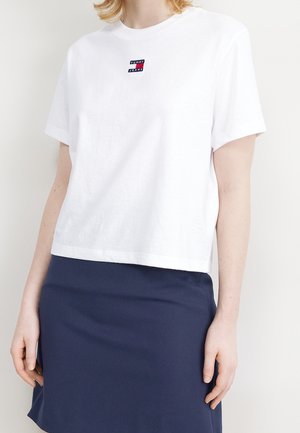 Kvinde iført en hvid Tommy Jeans kort t-shirt og en mørkeblå nederdel, stående med armene afslappet ved siden.