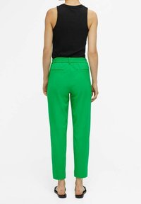 Pantalon vert taille haute avec ceinture élastique et poches arrière, associé à un haut noir ajusté sans manches et des sandales plates.