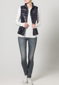 Mujer vistiendo un chaleco acolchado azul oscuro sobre una camiseta de manga larga blanca, con jeans azules y zapatillas blancas, de pie contra un fondo neutro.