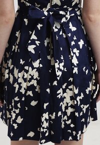 Marinblå klänning med vitt fjärilsmönster, veckad kjol och en tygbåge i midjan. Har en central dragkedjestängning bak.