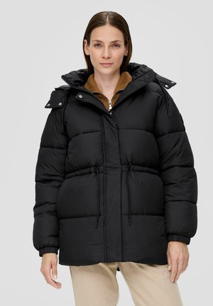 Frau in einer schwarzen gepolsterten Jacke mit Kapuze, darunter ein brauner Reißverschluss-Pullover, und beigen Hosen, stehend vor einem schlichten hellen Hintergrund.