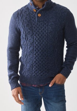 Homme portant un pull torsadé bleu marine avec des boutons en bois et un jean bleu, une main dans la poche et l’autre pendante.