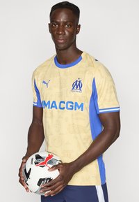 Ανοιχτό κίτρινο ποδοσφαιρικό μπλουζάκι με μπλε λεπτομέρειες, που διαθέτει υφή, λογότυπο ομάδας και έντονα μπλε γράμματα· συνοδεύεται από λευκή μπάλα ποδοσφαίρου.