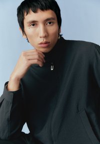 Sötét szürke cipzáras dzseki magas nyakkal, texturált anyagból, enyhe fényes hatással. A modellen rövid haj van, és az egyik kezét az állán pihenteti.