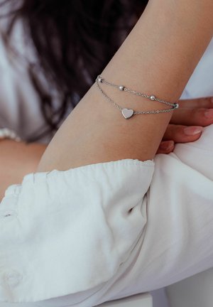 Silver double chain bracelet with heart charm and small beads worn on a wrist against a white blouse background.