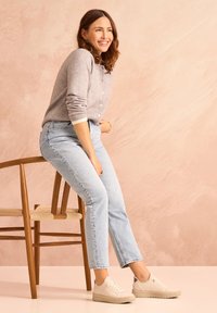 Gray knit cardigan, light blue jeans, and beige sneakers with silver accents, seated on a wooden chair against a soft pink background.