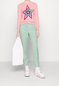 Suéter rosa con un diseño de estrella grande en colores llamativos, combinado con pantalones verdes con estampado y botas blancas de tobillo, llevando una tote bag blanca.
