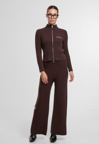 Hnědý žebrovaný pletený svetr s vysokým límcem a předním zipem, doplněný volnými hnědými pletenými kalhotami. Černá obuv na podpatku dokončuje vzhled.