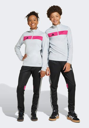 Zwei Kinder tragen Adidas-Sportbekleidung: hellgraue Zip-Jacken mit pinken Akzenten und schwarze Hosen mit pinken Details und weißen Streifen.