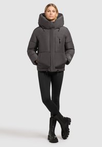 Grå puffjacka med stor huva, framdragkedja, två sidofickor och en dragkedjeficka på bröstet. Bärs med svarta leggings och stövlar.