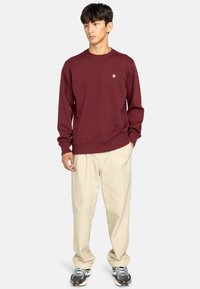 Burgunder sweatshirt med ribbet rund hals og manchetter, båret med beige bukser og grå sneakers. Har et lille logo på brystet.