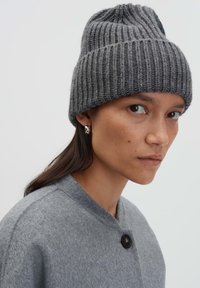 Una donna che indossa un berretto di maglia grigio, un orecchino a cerchio argentato e un cappotto di lana grigio con un bottone nero, che guarda leggermente di lato.