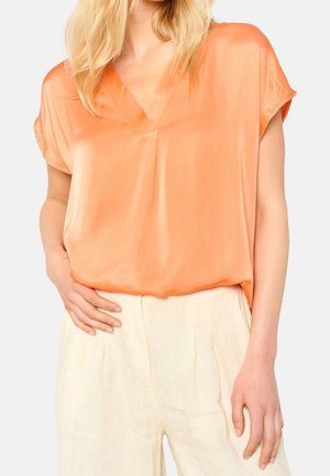Kobieta ubrana w luźną, pomarańczową satynową bluzkę z krótkim rękawem, wpuszczoną w kremowe lniane spodnie z wysokim stanem, stoi z ręką w kieszeni na jasnym tle.