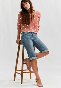Rosa prickig cardigan, blå denimshorts med uppvikt nederkant och ljusrosa klackar, sittande på en träpall mot en neutral bakgrund.