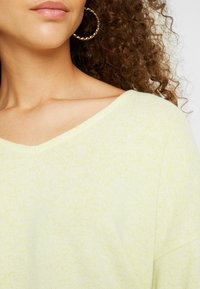 Jasnożółty sweter dziergany z dekoltem w kształcie litery V i teksturowanym materiałem. Częściowo widoczne kręcone włosy z dużą skręconą kolczykiem-kołem.