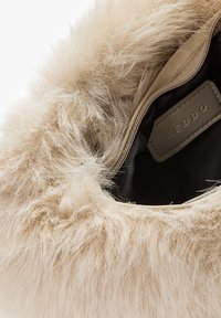 Beige čizmama obloženim krznom s mekom, pahuljastom teksturom. Ima tamnobraon kožni detalj i crni unutrašnjost, označen logotip oznakom.