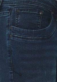 Primer plano de unos vaqueros de mezclilla azul oscuro que muestran el bolsillo delantero con remaches y costuras detalladas.